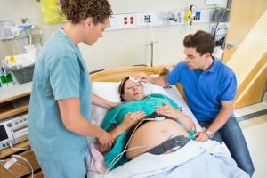 A pregnant patient lying in a bed, with a doctor and a loved one standing either side of them.