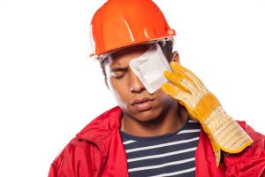 A construction worker wearing an orange helmet, holding his eye in pain. His eye has a white bandage over it. 