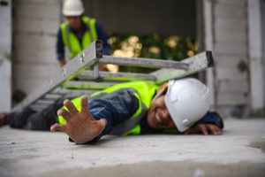 A worker in a hi-vis jacket and white safety helmet lies on the floor with a ladder lying on top of him.